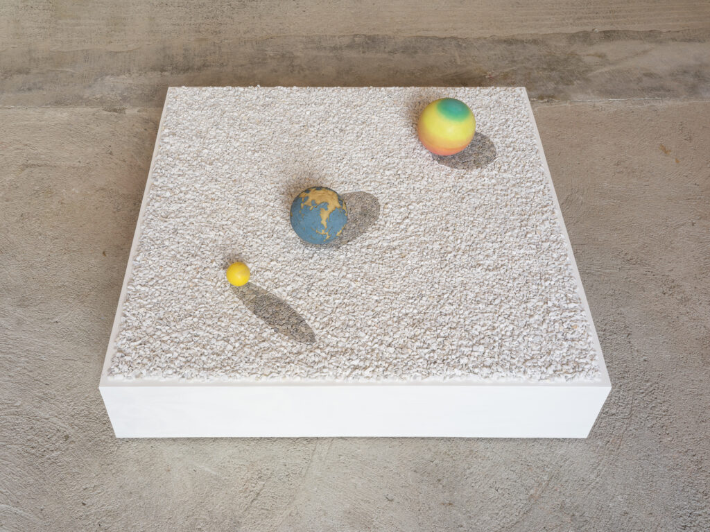 Installation view of an artwork consisting of a square plattform with white gravel and tree colored balls sitting on it.