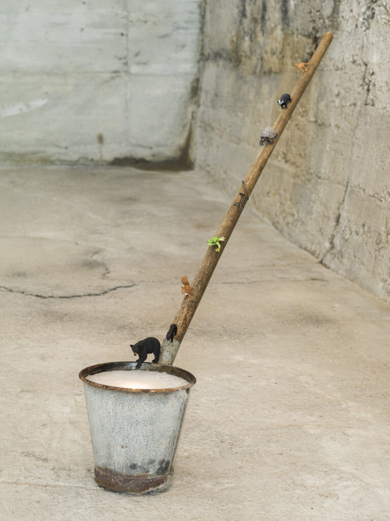 Installation view of an artwork consisting of a long-handeled dipper, animal figurins walking on the handle and the dipper containing a candle.